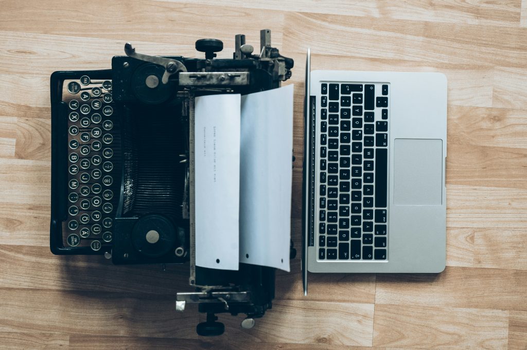 A typewriter and laptop placed back-to-back on a wooden surface.