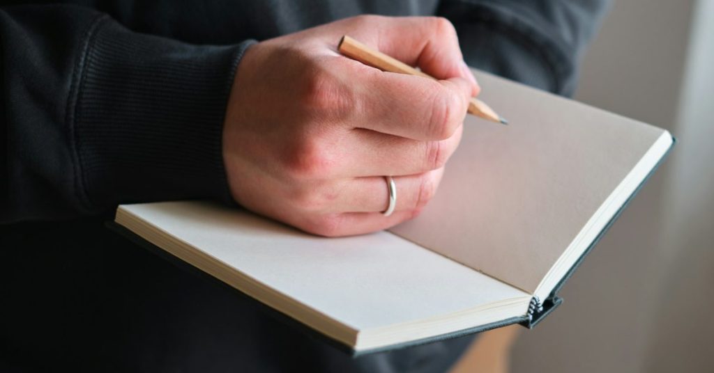 A hand holding a pencil over a blank notebook, ready to write something