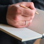 A hand holding a pencil over a blank notebook, ready to write something