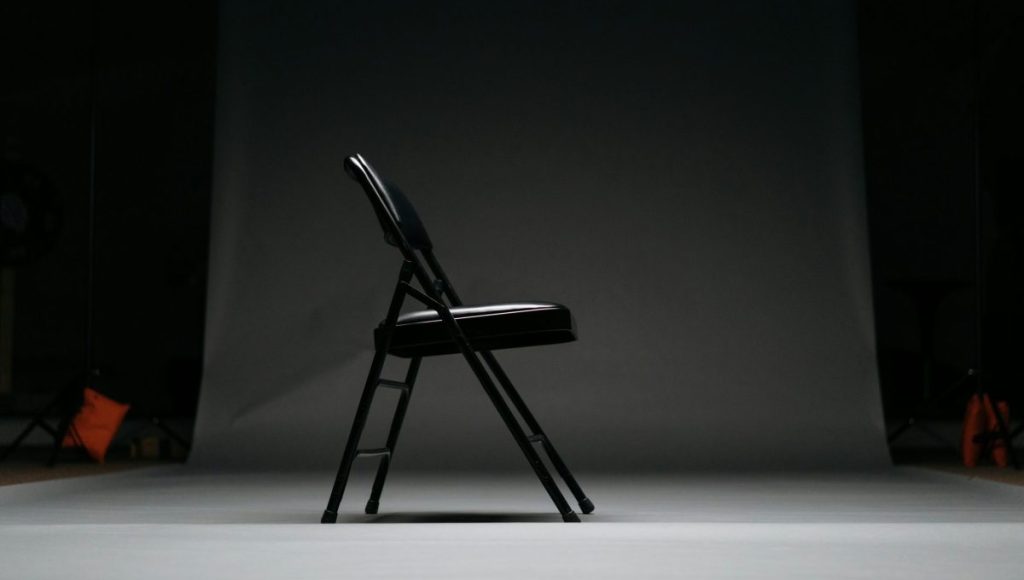 A black folding chair facing right in an empty room with no one sitting on it.