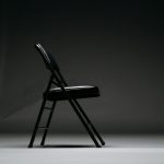 A black folding chair facing right in an empty room with no one sitting on it.