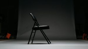 A black folding chair facing right in an empty room with no one sitting on it.