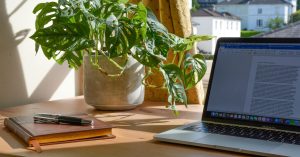 Wooden desk in front of the window with a laptop on the right and a notebook and pen on the left. Behind both is a potted plant.